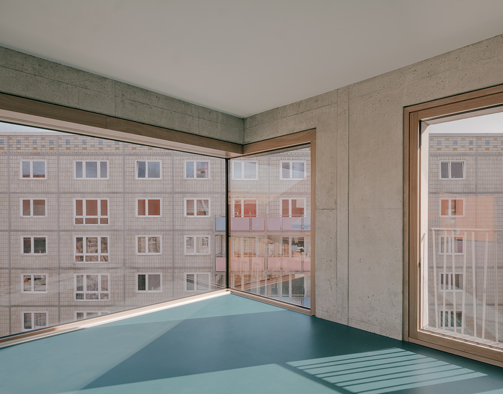Blick auf den Innenraum des Wohnhauses in der Magazinstraße in Berlin mit unbekleideter Leichtbetonaußenand, warmen Farbton der Fensterprofile, weißer Decke und Boden aus einer blauen Kunststoffbeschichtung