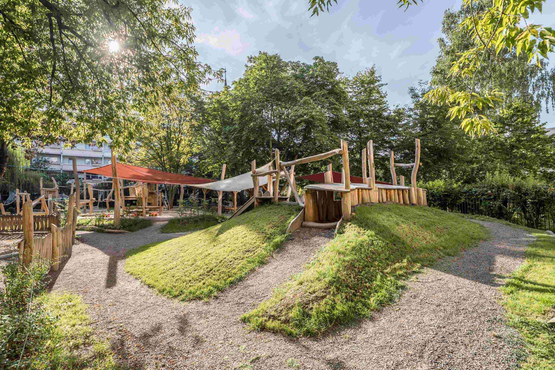 Ein Spielplatz im Freien in einer üppig grünen Umgebung mit Bäumen und Sonnenlicht, das durch das Laub filtert. Der Spielplatz umfasst Holzstrukturen wie ein Spielhaus, Brücken und Plattformen sowie grasbewachsene Hügel.